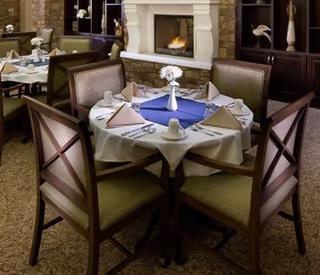 A dining area with a round table set for six people. The table is covered with a white tablecloth and a blue table runner, with folded napkins, cups, and a vase with white flowers in the center. In the background, there is a fireplace with a fire burning and dark wooden shelves with decorative items.