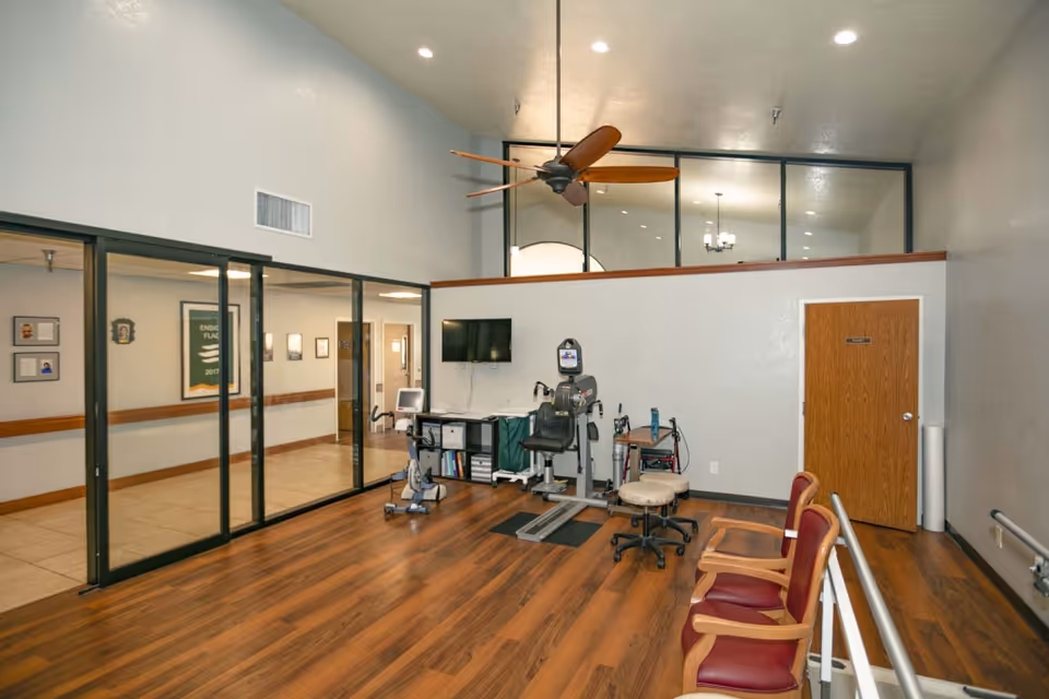 A rehabilitation or therapy room with wooden flooring, exercise equipment including a stationary bike and a seated exercise machine, a wall-mounted TV, and several chairs with wooden arms and red cushions. The room has a high ceiling with a ceiling fan and large glass windows looking into a hallway.