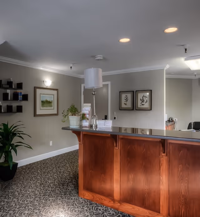 Reception area with a wooden front desk, a table lamp, potted plants, framed artwork on the walls, and patterned carpet flooring in a memory care community facility.