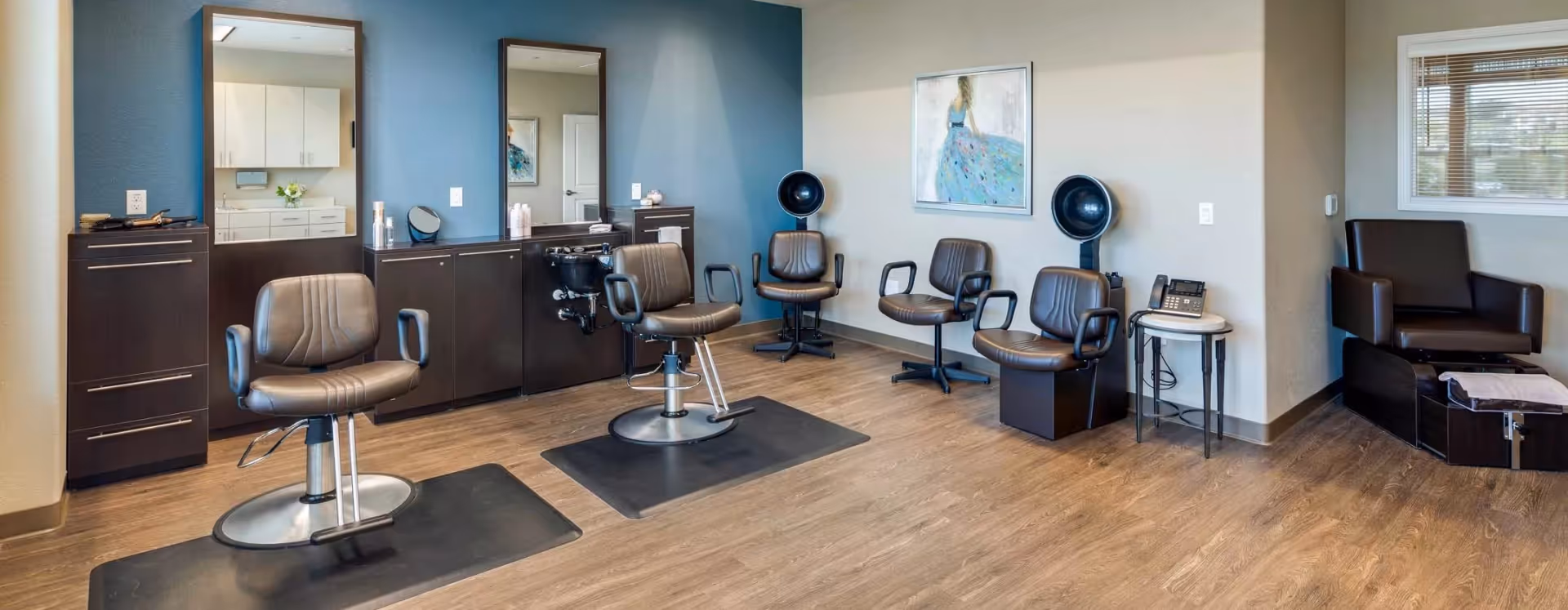 Interior view of a salon area in a senior living facility with two styling chairs in front of mirrors and cabinets, three waiting chairs, a hair dryer, and a pedicure station with a chair and foot bath. The room has wood flooring and neutral-colored walls with a framed artwork.
