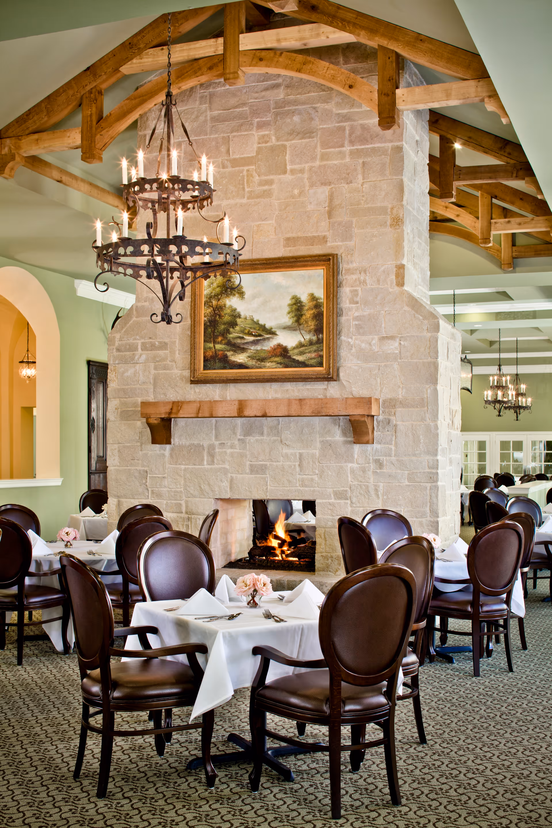 Elegant dining room with round tables covered in white tablecloths, set with napkins and silverware. Dark wooden chairs with leather seats surround the tables. A large stone fireplace with a lit fire is the focal point, above which hangs a framed landscape painting. The ceiling features exposed wooden beams and a wrought iron chandelier with lit candles.