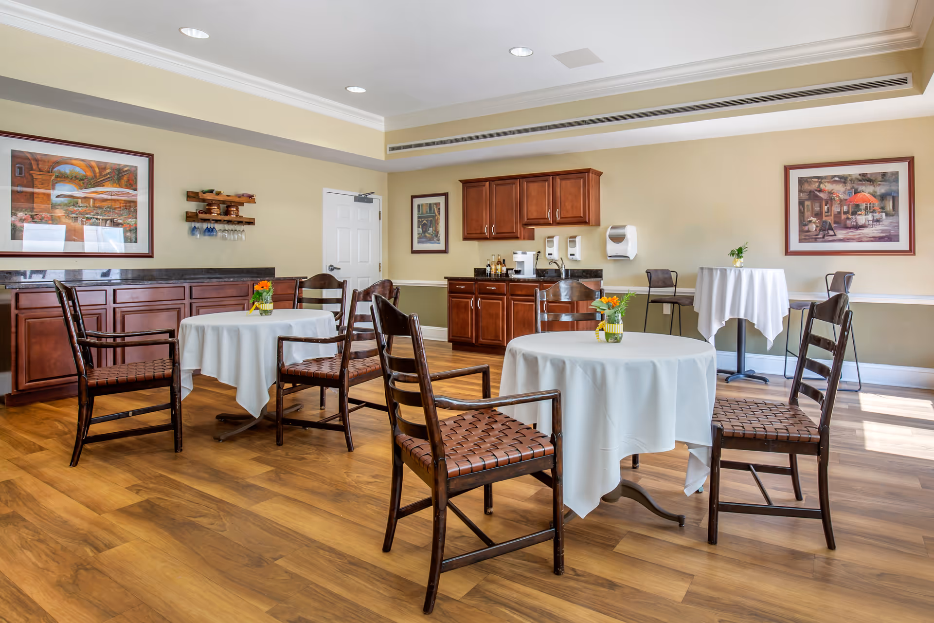 A bright dining room with wooden floors and several round tables covered with white tablecloths. Each table is surrounded by wooden chairs with woven seats. The room has beige walls adorned with framed artwork and features wooden cabinets and a countertop with a coffee maker and condiments. There are also two high tables with chairs near the wall.