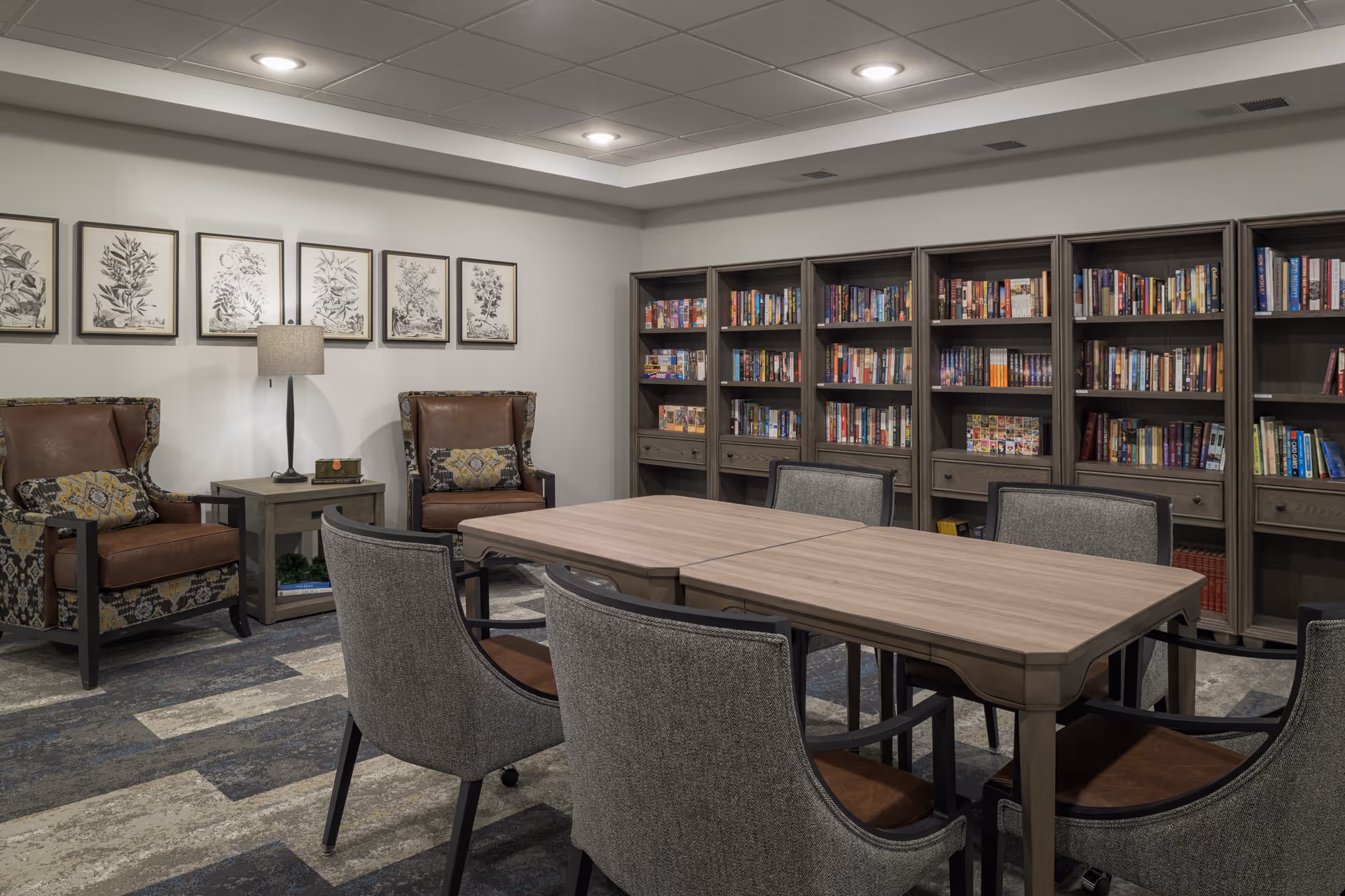 A cozy reading or meeting room with a wooden table surrounded by six upholstered chairs. Against the wall are several bookshelves filled with books. Two leather armchairs with patterned cushions flank a small side table with a lamp. Botanical artwork is displayed on the wall above the armchairs.