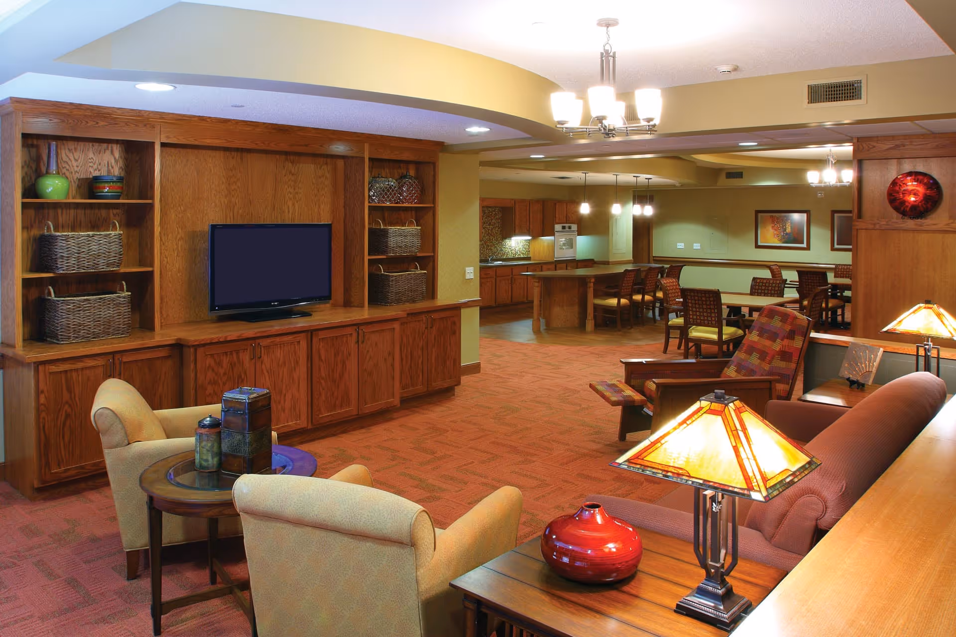 Open communal living area with seating, a built-in wooden entertainment center and TV, lamps, and a dining/kitchen area in the background.
