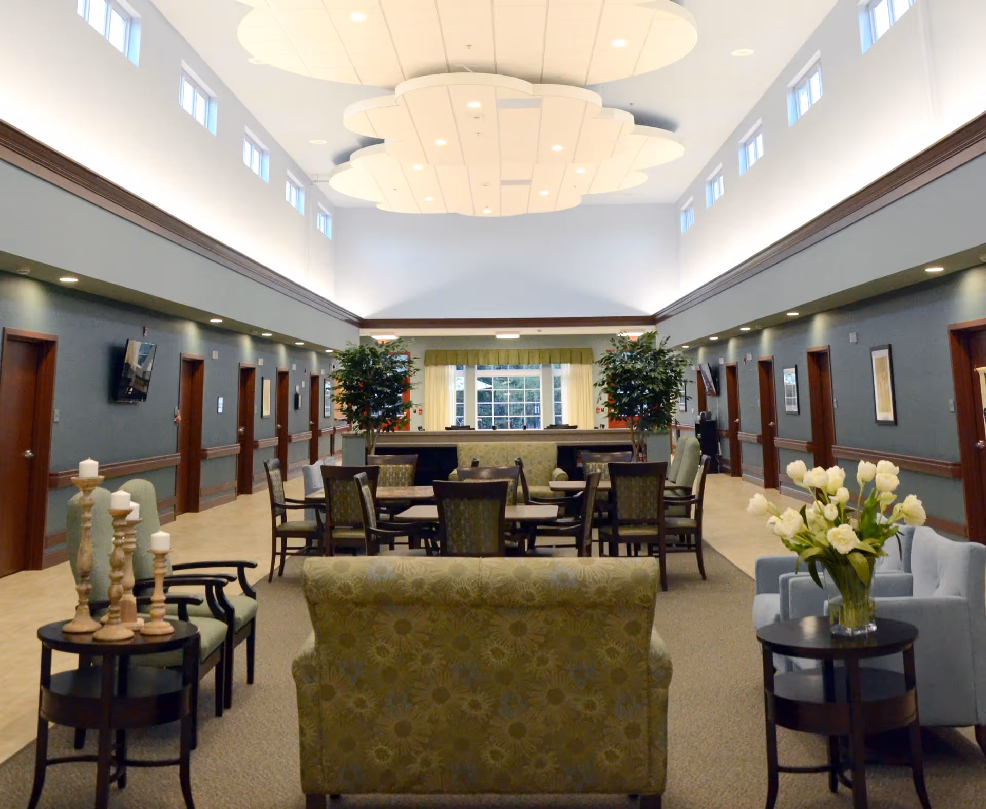 A spacious and well-lit common area in a senior living facility with high ceilings and multiple small windows near the ceiling. The room features several seating arrangements including armchairs, sofas, and tables with chairs. There are decorative plants and flowers placed around the room, and the walls have framed artwork and mounted televisions. The area has a calm and welcoming atmosphere.