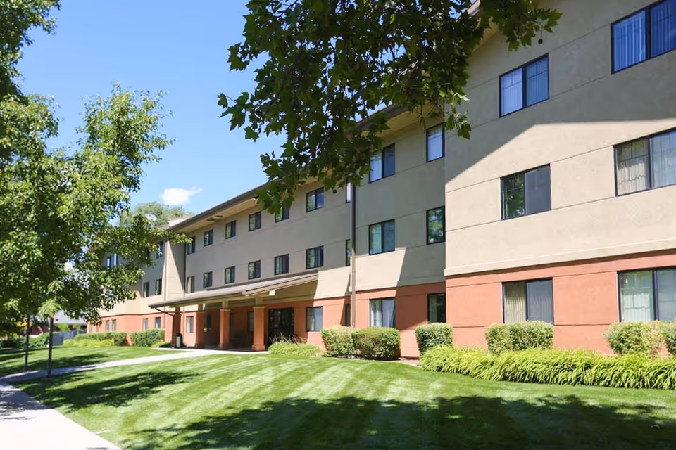 Exterior view of a three-story senior living facility building with beige and reddish-brown walls, multiple windows, a covered entrance, well-maintained green lawn, bushes, and trees under a clear blue sky.
