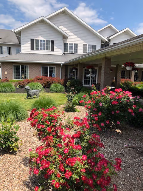 Exterior view of a senior living facility building with white siding and brick accents. The foreground features a garden with vibrant red flowers, green shrubs, and a decorative dog statue on the lawn. The sky is partly cloudy with blue patches visible.