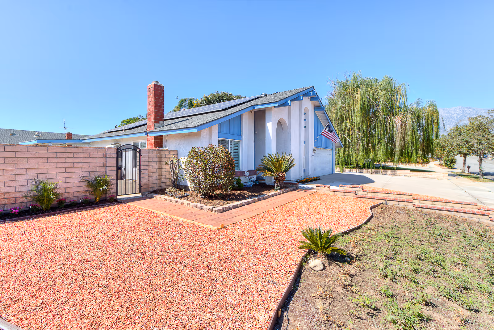 Exterior view of a single-story house with a light-colored facade and blue trim. The front yard features red gravel landscaping, a small garden area with plants, a brick chimney, and an American flag mounted near the garage. A clear blue sky is visible above.