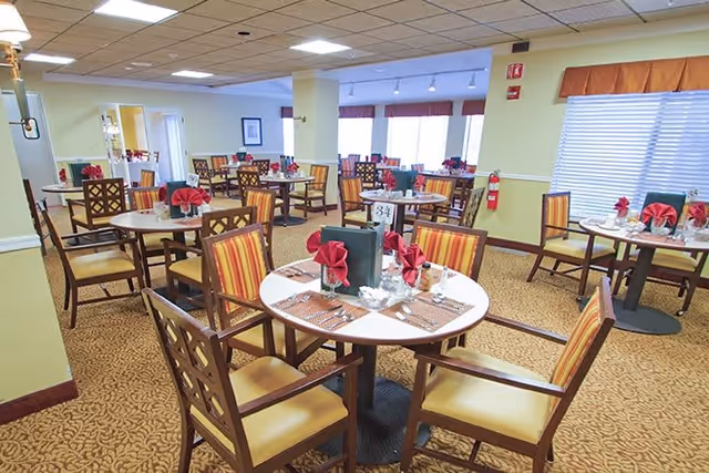 A well-lit dining room with multiple round tables set with placemats, napkins folded in a decorative style, glasses, and menus. The room has patterned carpet flooring, wooden chairs with striped cushions, and windows with blinds and valances. The walls are painted light yellow with white trim, and there is a fire extinguisher mounted on one wall.