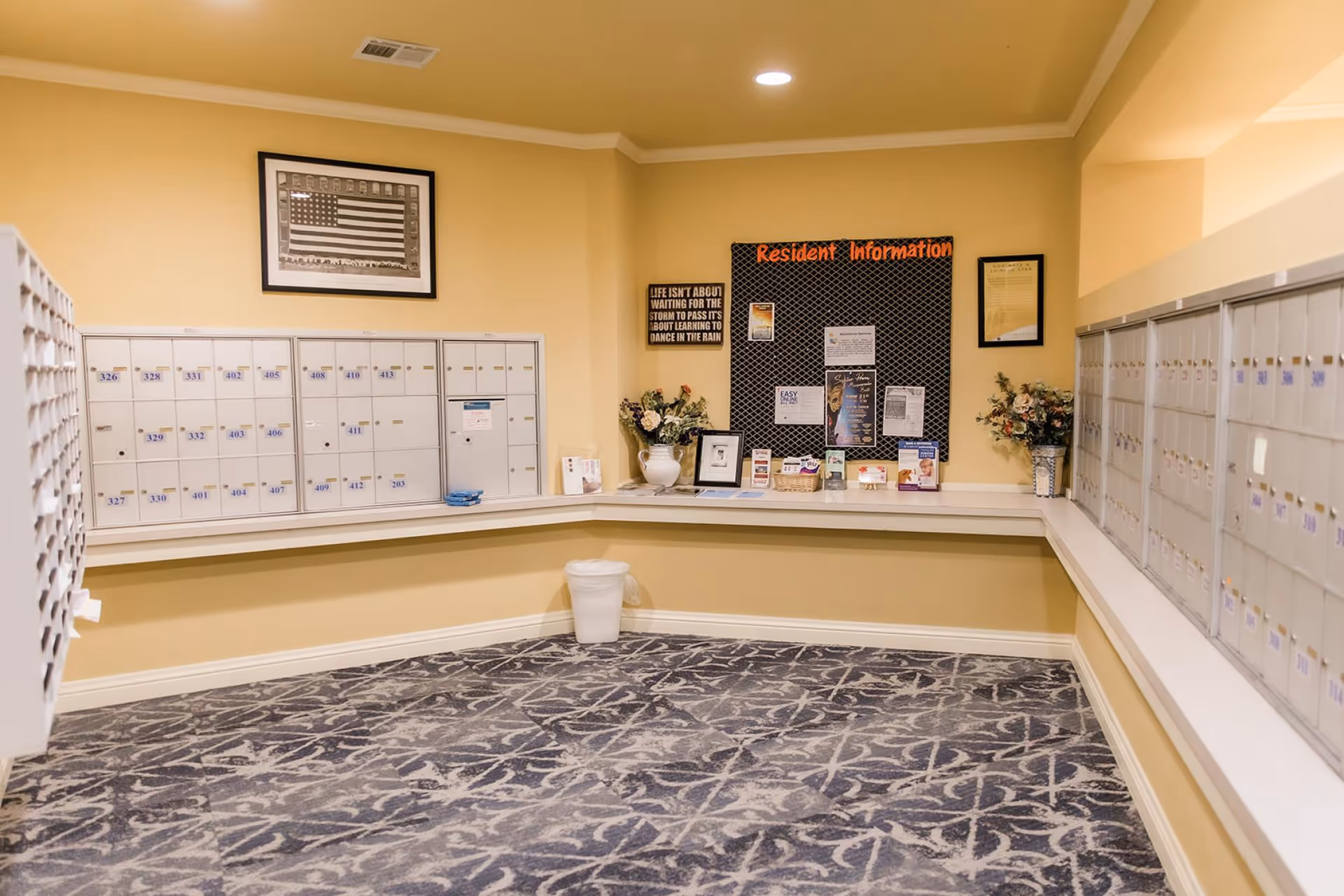 Mailroom with rows of numbered mailboxes, a resident information bulletin board, and decorative flowers.