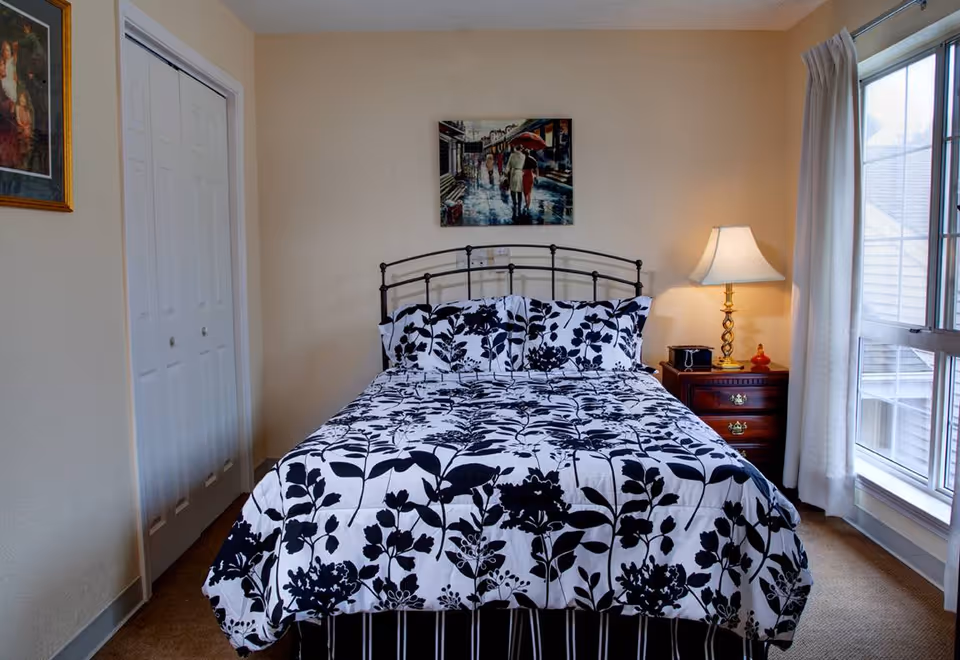 Bedroom with a metal-frame bed dressed in black-and-white floral bedding, a wooden nightstand with a lamp, closet doors and a large window.