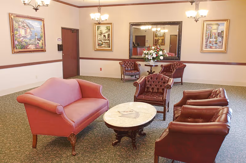 A sitting area in a care center with a pink upholstered loveseat, three brown leather armchairs, and a round marble-top coffee table. The room has beige walls with a brown chair rail, three framed paintings, a large mirror, and two chandeliers. A small table with a floral arrangement is placed against the wall.