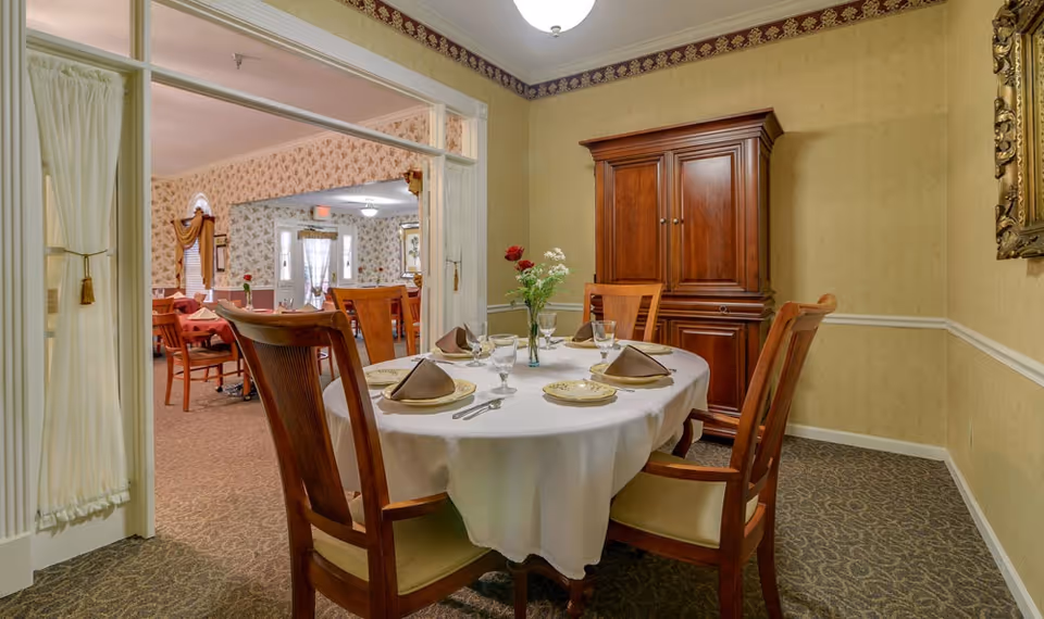 A dining room in a senior living facility with a round table covered with a white tablecloth set for four people. The table has plates, glasses, silverware, and folded brown napkins. A small vase with flowers is in the center. The room has beige patterned wallpaper, a wooden cabinet against the wall, and a framed mirror. Through an open doorway, another dining area with tables and chairs is visible.