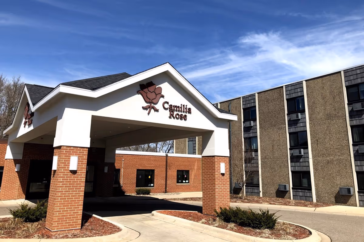 Exterior view of Camilia Rose Care Center showing the entrance canopy with a rose logo and the facility name. The building has a brick and concrete facade with multiple windows and air conditioning units. The sky is clear with some clouds.
