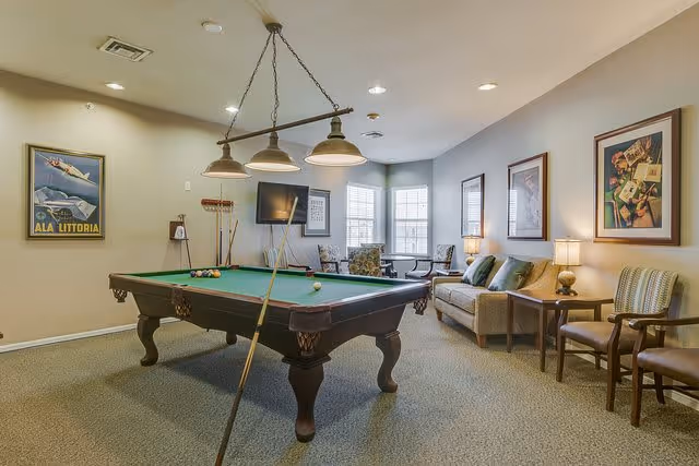 A cozy game room with a pool table in the center, a cue stick resting on the table, and a set of billiard balls arranged. The room has beige walls adorned with framed artwork, a flat-screen TV mounted on the wall, a sofa with cushions, two armchairs, and a side table with a lamp. Large windows allow natural light to brighten the space.
