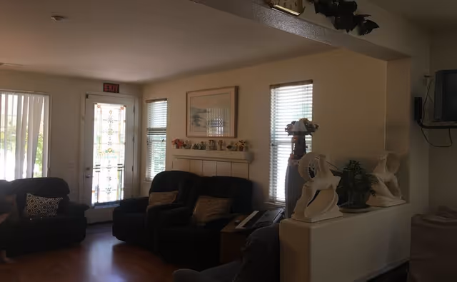 A cozy living room area with dark cushioned chairs and a couch, a decorative shelf with small figurines and a framed picture on the wall, two windows with blinds, a glass door with a stained glass design, and decorative sculptures on a half wall. The room has wooden flooring and soft lighting.