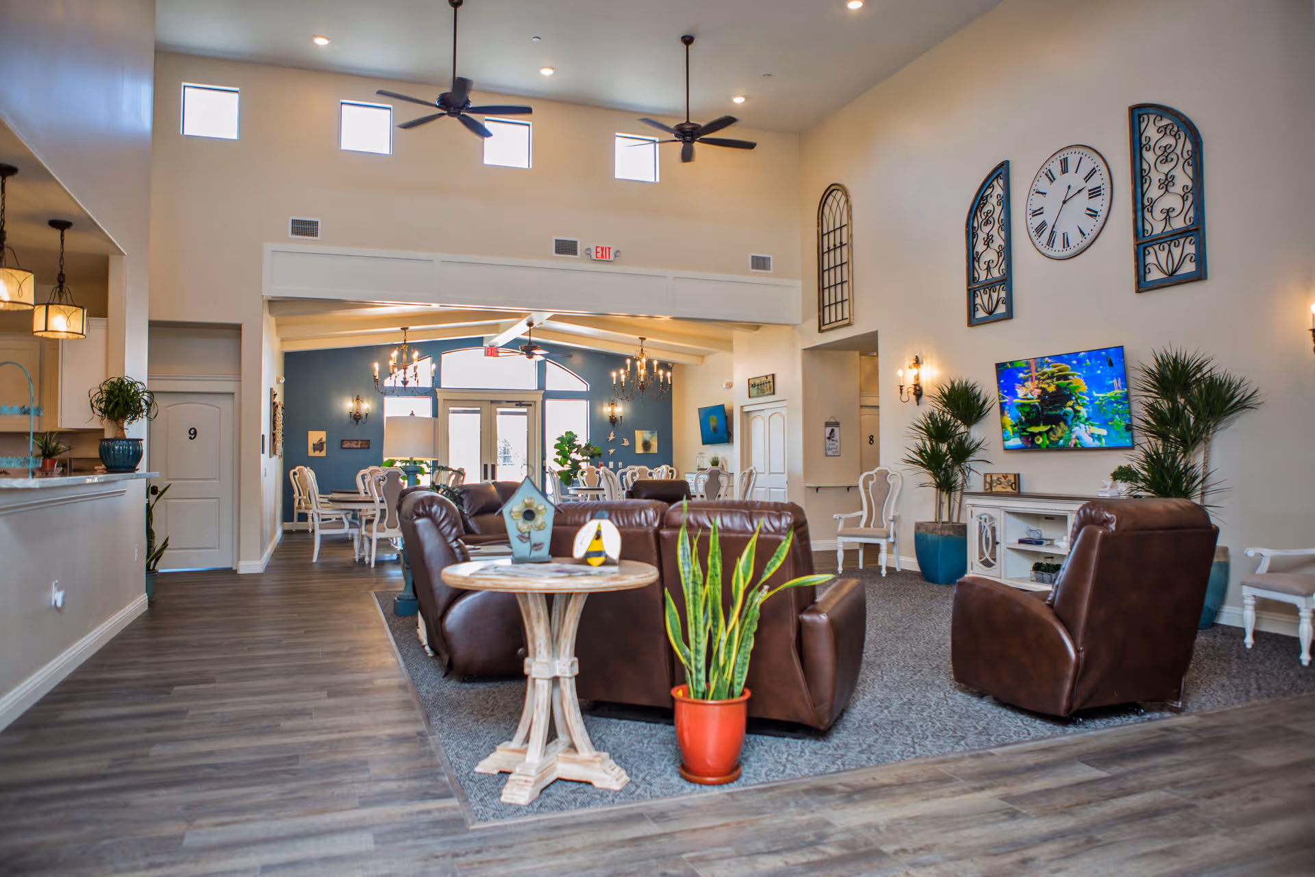 A spacious and well-lit living room area in an assisted living facility featuring brown leather recliners arranged around a small wooden table with decorative items and a potted plant. The room has high ceilings with ceiling fans, wall-mounted TV displaying an aquarium scene, decorative wall art including a large clock, and several potted plants. In the background, there is a dining area with white chairs and tables, chandeliers, and large windows letting in natural light.