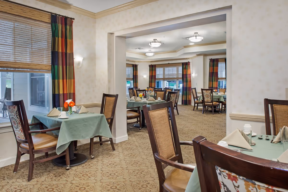 Interior view of a dining room in a senior living facility with tables covered in green tablecloths, set with napkins, glasses, and small flower vases. The room has patterned carpet, wooden chairs with woven backs and floral upholstery, large windows with bamboo blinds and colorful striped curtains, and ceiling lights.