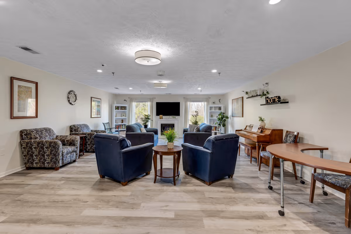 A spacious and well-lit living room in a senior living facility featuring multiple armchairs arranged around a small round table with a plant. The room has light-colored walls, wood flooring, a piano against one wall, a TV mounted above a fireplace, and large windows with white curtains letting in natural light. Shelves with books and decorative items are also visible.
