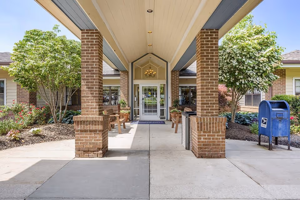 Covered brick entryway with a porte-cochère leading to glass double doors, benches, landscaping, and a blue mailbox.