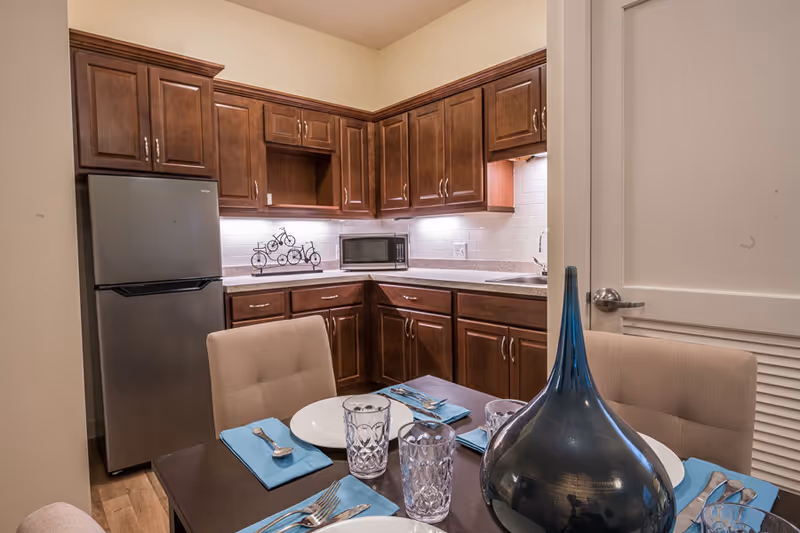 Kitchenette with dark wood cabinetry, a stainless refrigerator and microwave, and a dining table set with plates, blue napkins and a large decorative vase.