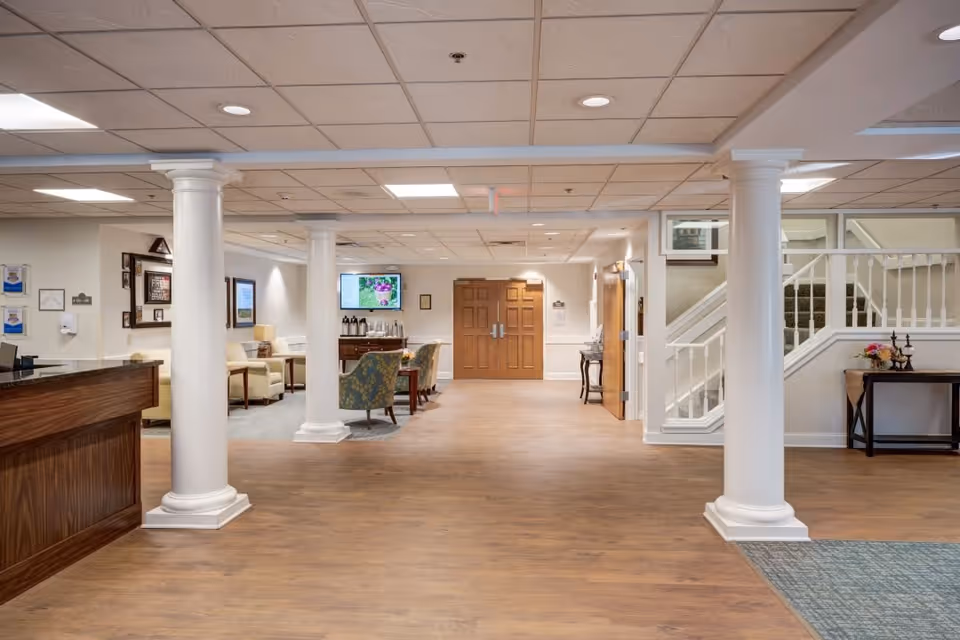 Interior view of a senior living facility lobby with wooden flooring, white columns, a reception desk on the left, seating area with chairs and a table in the background, a TV mounted on the wall, and a staircase on the right side.