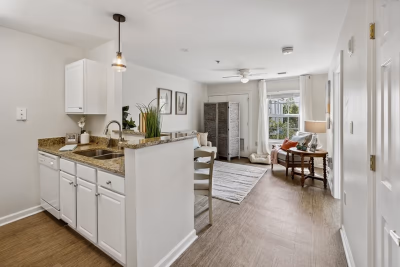 A bright and clean interior space featuring a small kitchen area with white cabinets, granite countertops, and a sink. Adjacent to the kitchen is a living area with a light-colored rug, a small sofa, a wooden side table with a lamp, and a window with white curtains letting in natural light. The floor is wood-style, and the walls are painted white. There is a decorative plant on the kitchen counter and framed artwork on the wall.