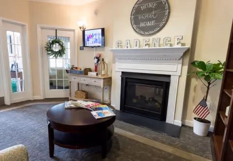 A cozy living room area with a round wooden coffee table holding magazines, a white fireplace with the word 'CADENCE' displayed on the mantel, a round wall decoration above the fireplace that says 'Home sweet Home', a small table with decorative items and a TV mounted on the wall, a potted plant with an American flag, and glass doors with a wreath hanging on one.