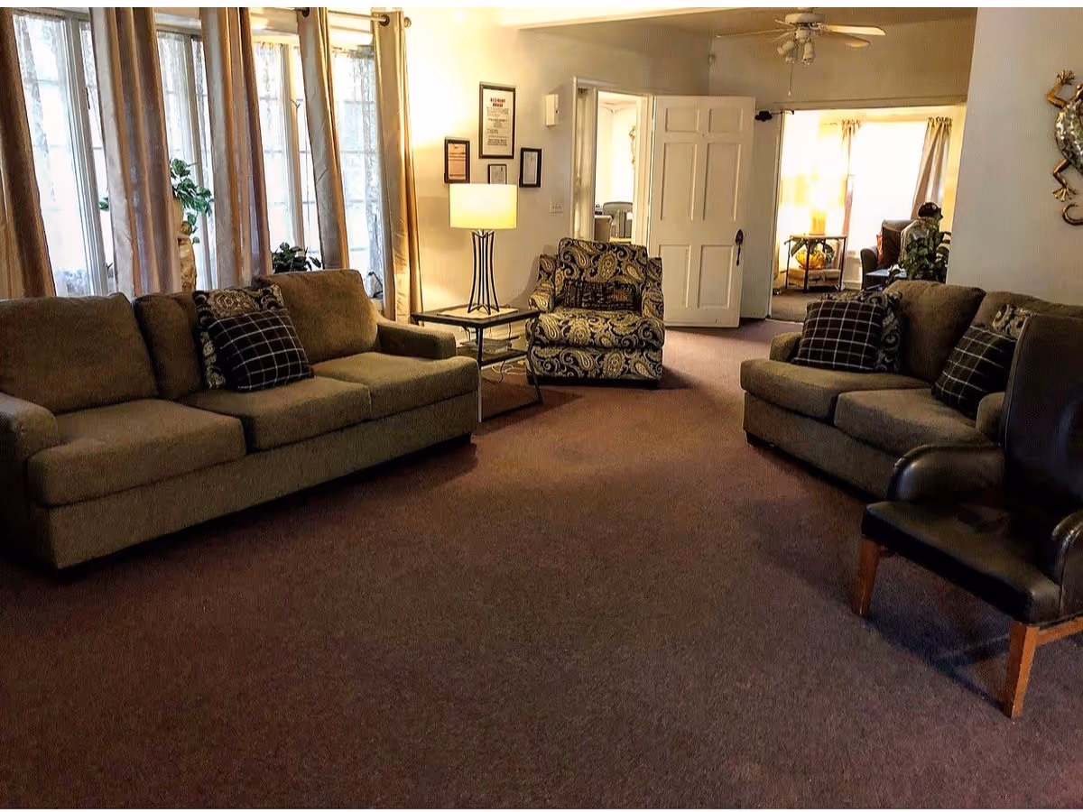 A cozy living room with two beige sofas adorned with patterned and checkered pillows, a patterned armchair, a black leather chair, a side table with a lamp, and large windows with curtains allowing natural light to fill the space.