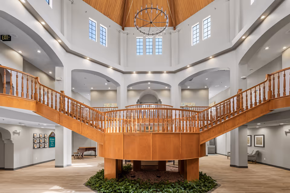 Spacious senior living community interior with a large wooden staircase and railing leading to an upper level. The area features high ceilings with multiple windows allowing natural light, a chandelier hanging from a wooden ceiling, and plants arranged below the staircase. The walls are painted light gray, and there are framed pictures and seating areas visible in the background.