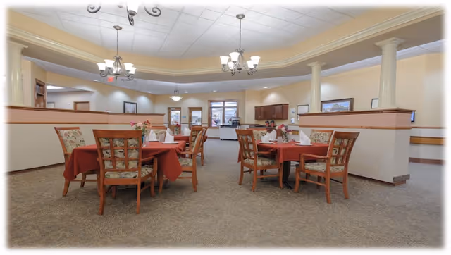 Interior view of a senior living facility dining room with several tables covered in red tablecloths, each set with napkins and floral centerpieces. The room features beige walls, carpeted floors, decorative columns, and ceiling light fixtures.