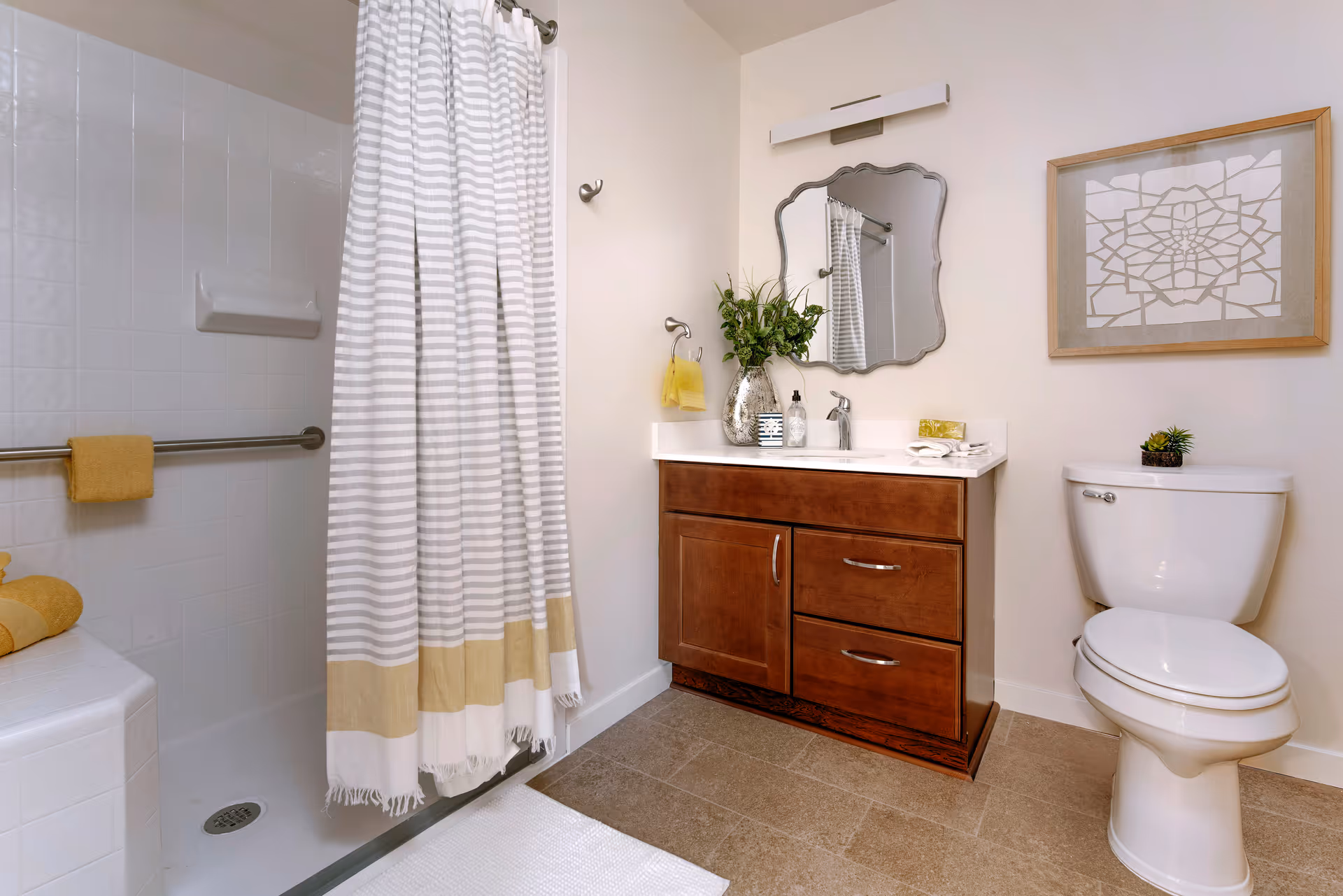 A clean and well-lit bathroom featuring a shower with a white and beige striped curtain, a wooden vanity with a white countertop, a decorative mirror above the sink, a toilet, and a framed geometric artwork on the wall. There are yellow towels and a small plant on the toilet tank.