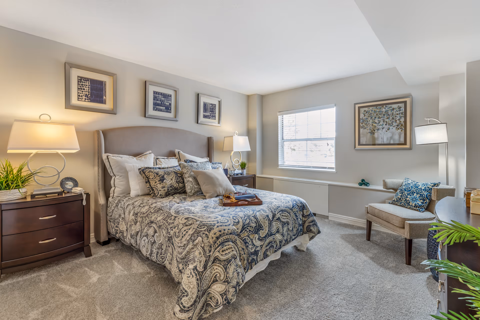 A well-lit bedroom with a large bed featuring a patterned blue and beige comforter and multiple pillows. There are two dark wooden nightstands on either side of the bed, each with a modern lamp and decorative items. Above the bed are three framed abstract artworks. A window with blinds lets in natural light, and a beige armchair with a blue patterned pillow sits near the window. The room has light-colored walls, carpeted floor, and additional framed artwork on the wall beside the window.