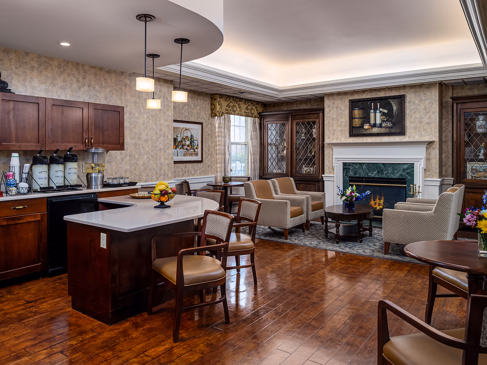 A cozy common area in a senior living facility featuring a small kitchen island with chairs, a countertop with beverage dispensers, and a seating area with armchairs arranged around a coffee table in front of a fireplace. The room has wooden floors, warm lighting, and decorative cabinets and artwork on the walls.