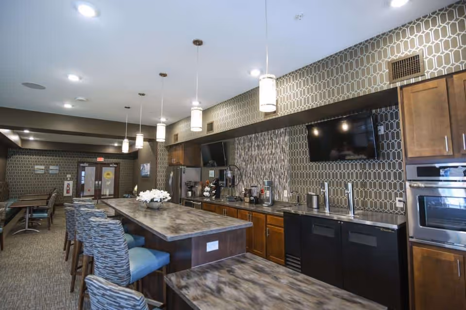 Interior view of a modern kitchen and dining area in a senior living facility. The space features a long island with patterned cushioned chairs, pendant lights hanging from the ceiling, wooden cabinets, stainless steel appliances including an oven and refrigerator, a mounted flat-screen TV, and a decorative backsplash. In the background, there is a seating area with tables and chairs near double doors.