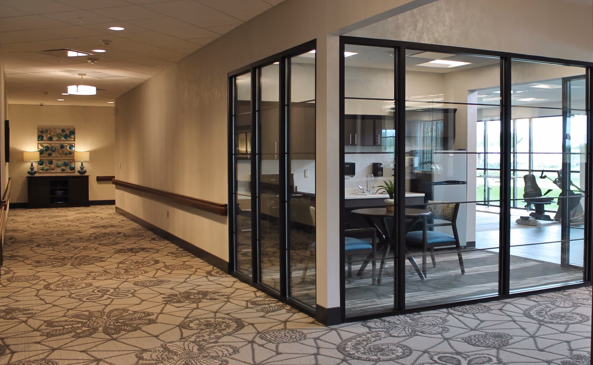 A hallway in a senior living facility with patterned carpet and beige walls. On the right side, there is a glass-walled room containing a small round table with chairs and a kitchenette area. In the background, exercise equipment is visible through the glass walls. The hallway is softly lit with ceiling lights and has a decorative console table with lamps and artwork at the end.