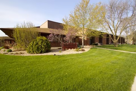 Brick senior living facility exterior with green lawn, trees, and curved walkway.
