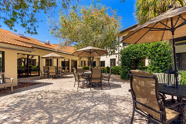 Sunny paved courtyard with patio tables and umbrellas beside a two-story building and landscaping.