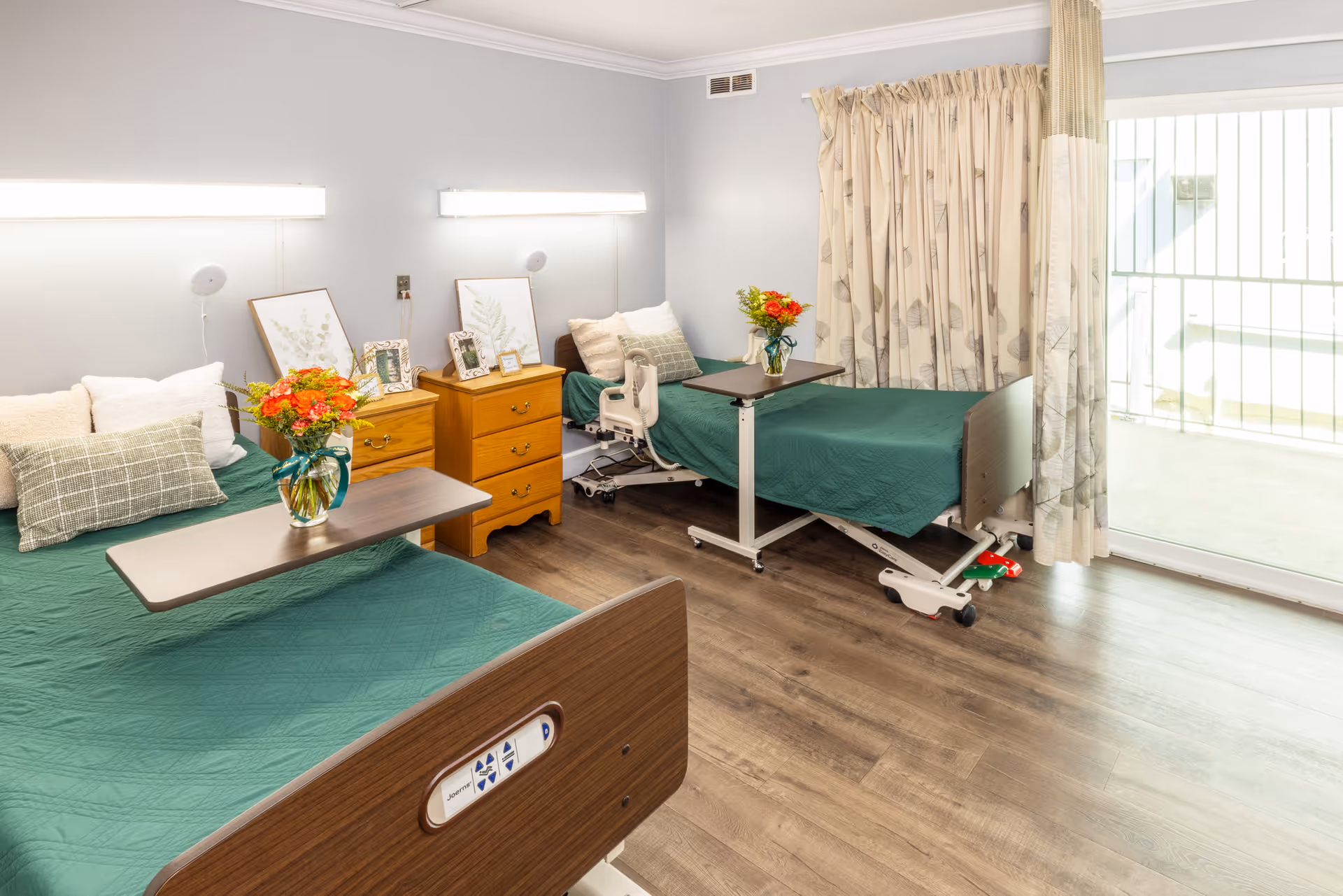 Shared patient room with two adjustable hospital beds with green bedspreads, bedside dressers, flowers on overbed tables, and a sliding glass door to a balcony.