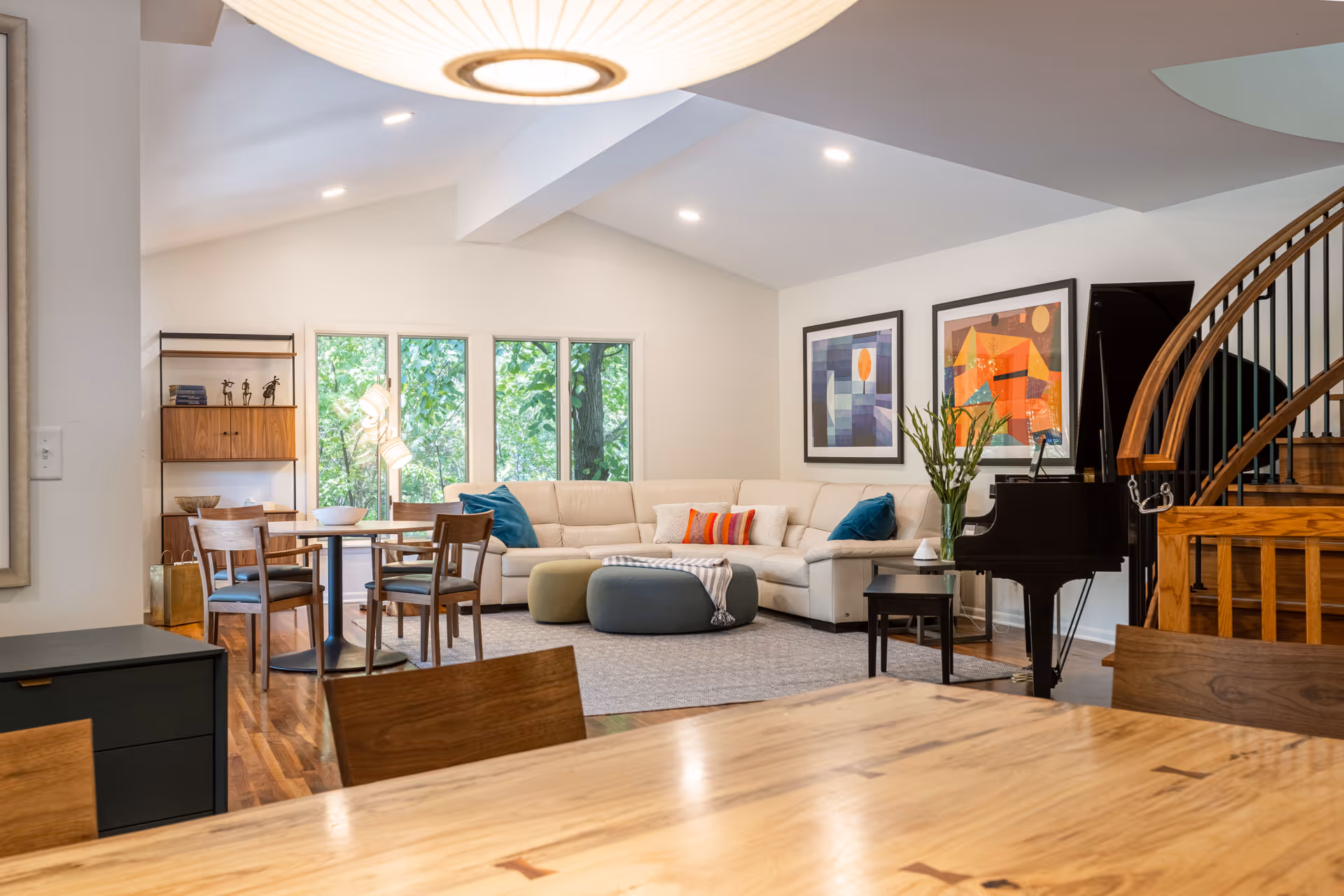 Bright modern living room with a large sectional sofa, dining table, grand piano and windows overlooking trees.