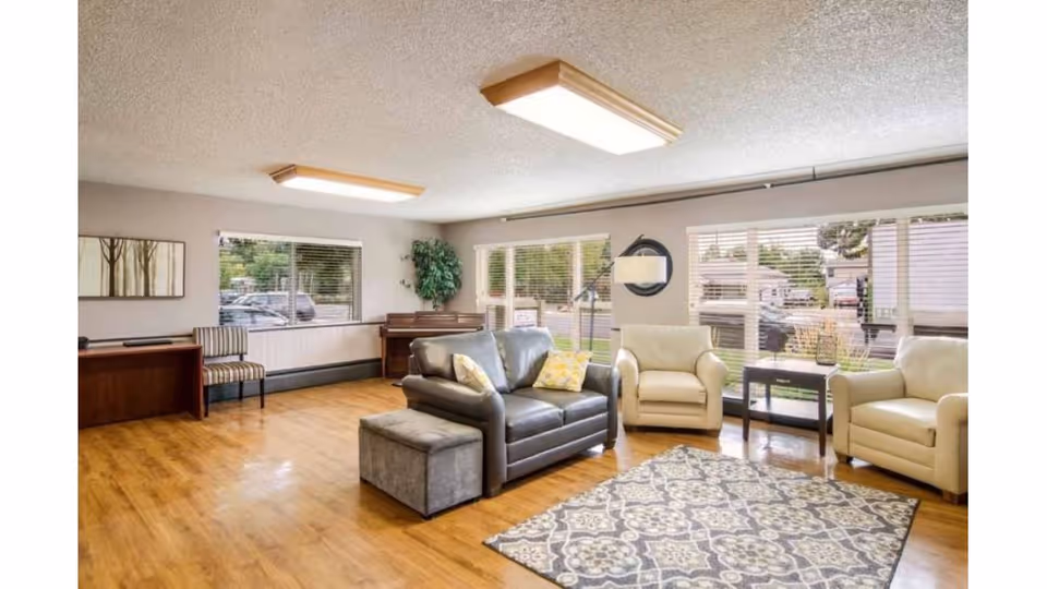 A bright and spacious living room with large windows letting in natural light. The room features a dark leather loveseat with two pillows, two cream-colored armchairs, a patterned area rug, a small side table, a floor lamp, a piano in the corner, and a wooden desk with a striped chair. The floor is wooden, and there is a framed artwork on the wall.