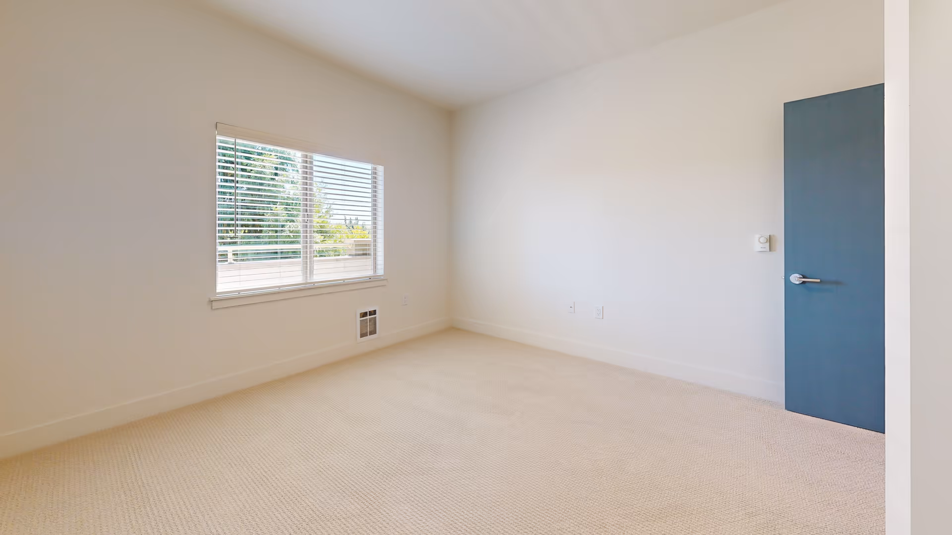 Empty room with beige carpet, white walls, a window with white blinds showing greenery outside, and a closed blue door on the right side.