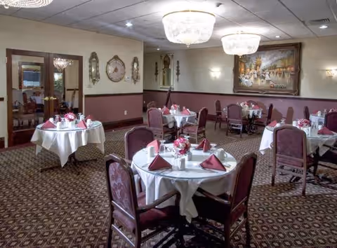 A dining room with round tables covered in white tablecloths, each set with folded burgundy napkins, plates, cups, and floral centerpieces. The room has patterned carpet, upholstered chairs, chandeliers, wall sconces, decorative mirrors, and a large framed painting on the wall.