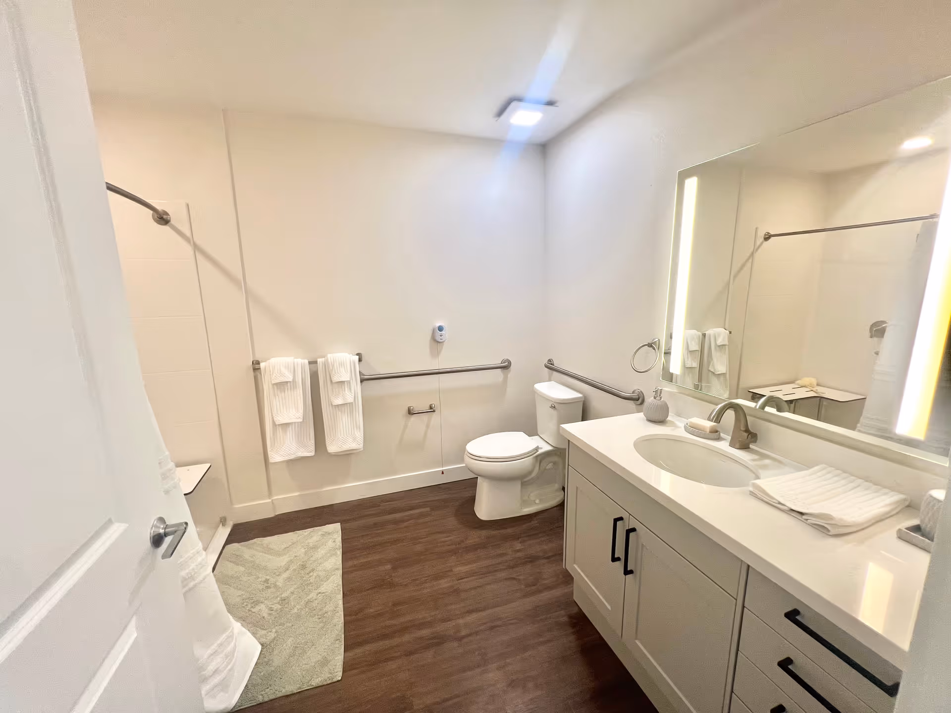 Accessible bathroom with a toilet, sink vanity and illuminated mirror, shower with curtain, grab bars and towels.