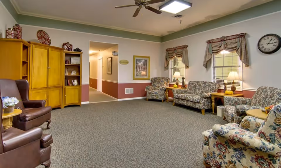 A cozy living room area in a senior living facility with floral patterned armchairs and a loveseat, two brown leather armchairs, wooden side tables with lamps, a wooden cabinet with decorative items, and two windows with valance curtains. The walls are painted white with a green trim near the ceiling and a reddish-brown lower half. A ceiling fan and a wall clock are also visible.