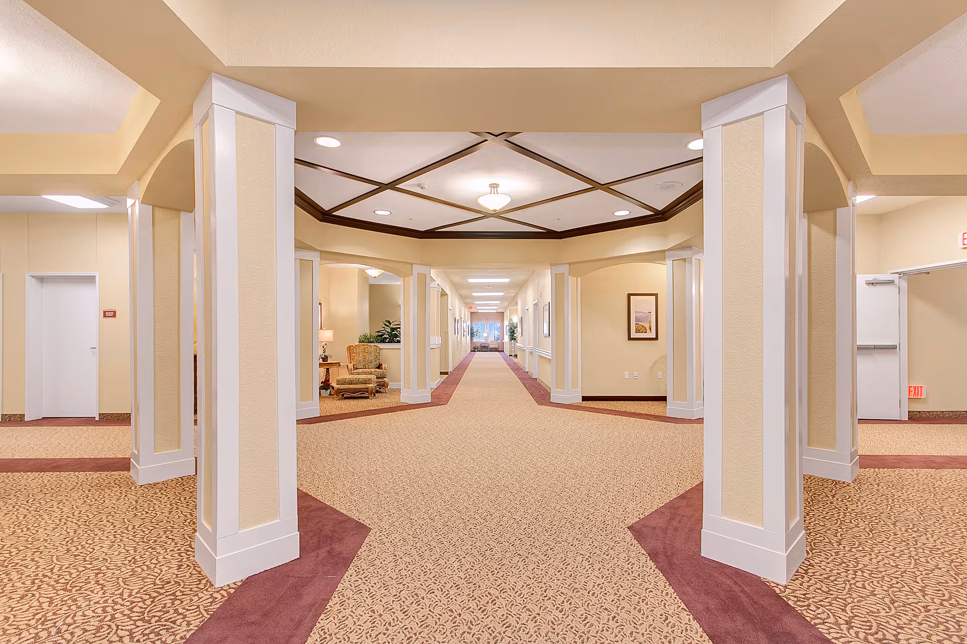 Wide hallway in a senior living facility with beige walls and patterned carpet. The hallway features white pillars, recessed ceiling lights, and a decorative ceiling design. There is a seating area with an armchair and lamp on the left side, and framed artwork on the walls. Doors and exit signs are visible at the far ends of the hallway.