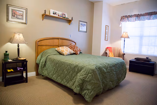 A cozy bedroom with a bed covered in a green quilt and decorative pillows. There is a wooden headboard, a nightstand with a lamp and some items, a framed picture on the wall, a small shelf with decorative items above the bed, a striped armchair with a throw blanket, a floor lamp, a window with blinds and a valance, and a dark ottoman near the window.