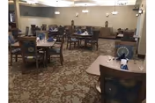 A spacious dining room with multiple wooden tables and chairs arranged neatly. Each table is set with blue napkins and silverware. The room has patterned carpet flooring, beige walls, and several chandeliers hanging from the ceiling providing warm lighting.