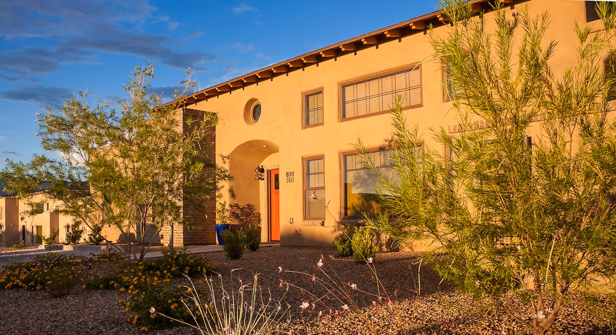 Exterior view of a southwestern-style building with tan stucco walls, a red door, and several windows. The foreground features desert landscaping with gravel, small bushes, and trees under a partly cloudy blue sky.