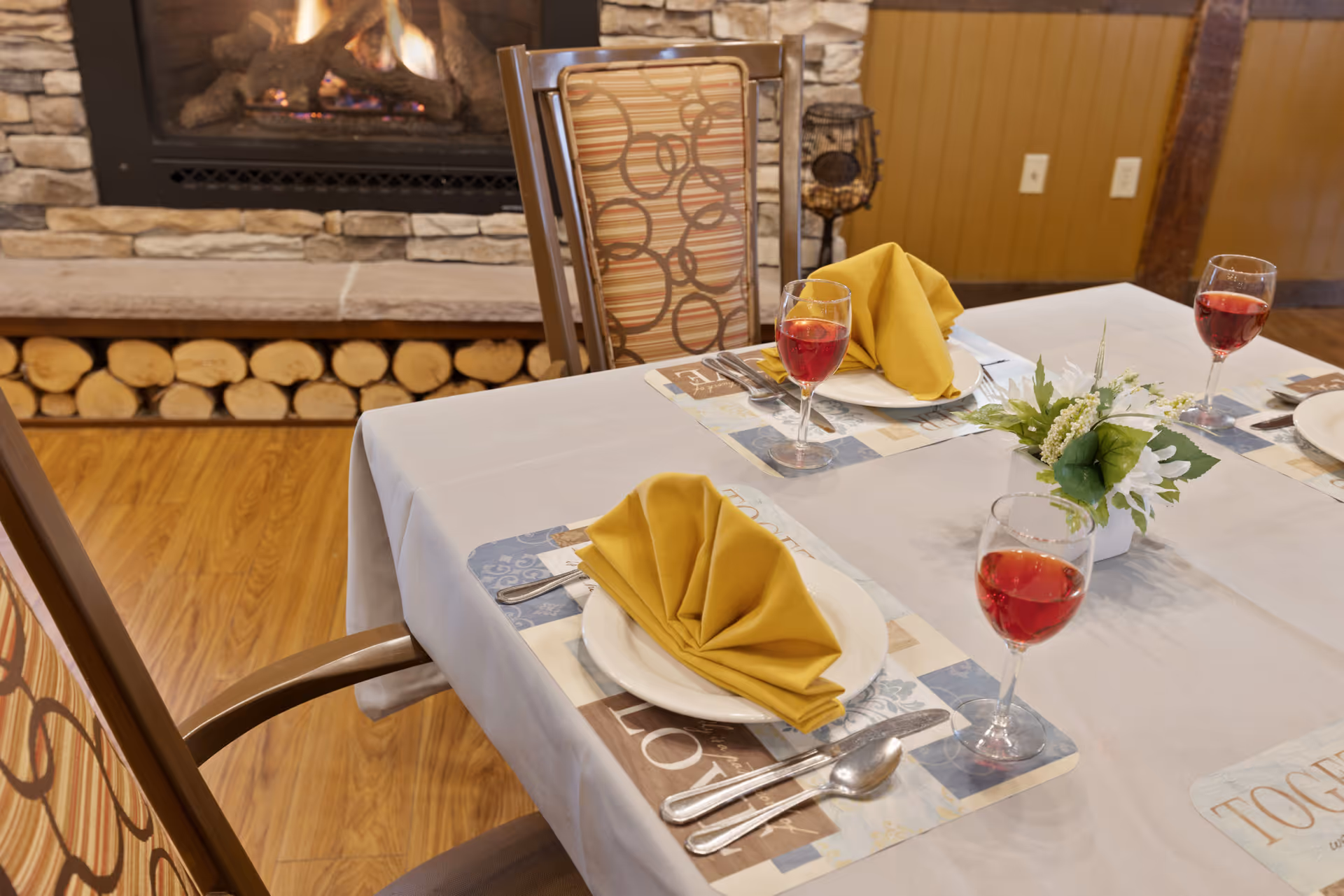 A dining table set for three with white tablecloth, yellow folded napkins on white plates, silverware, and glasses filled with red beverage. A small floral centerpiece is placed on the table. In the background, there is a stone fireplace with logs underneath and wooden flooring.